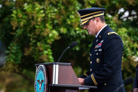 Man in Army dark blue service uniform is looking down at a podium while reading. The dark brown podium has the light blue round seal of the Department of Defense on the front. There are dark green trees in the background.
