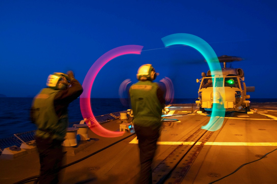 A sailor stands next to a fellow sailor as they wave red and green batons at an aircraft aboard a ship at sea.