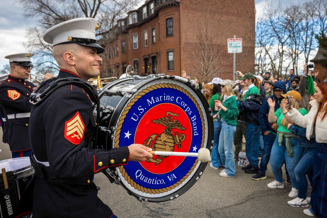 A Marine plays the drum as another plays the trumpet in front of a crowd dressed in green near row houses.