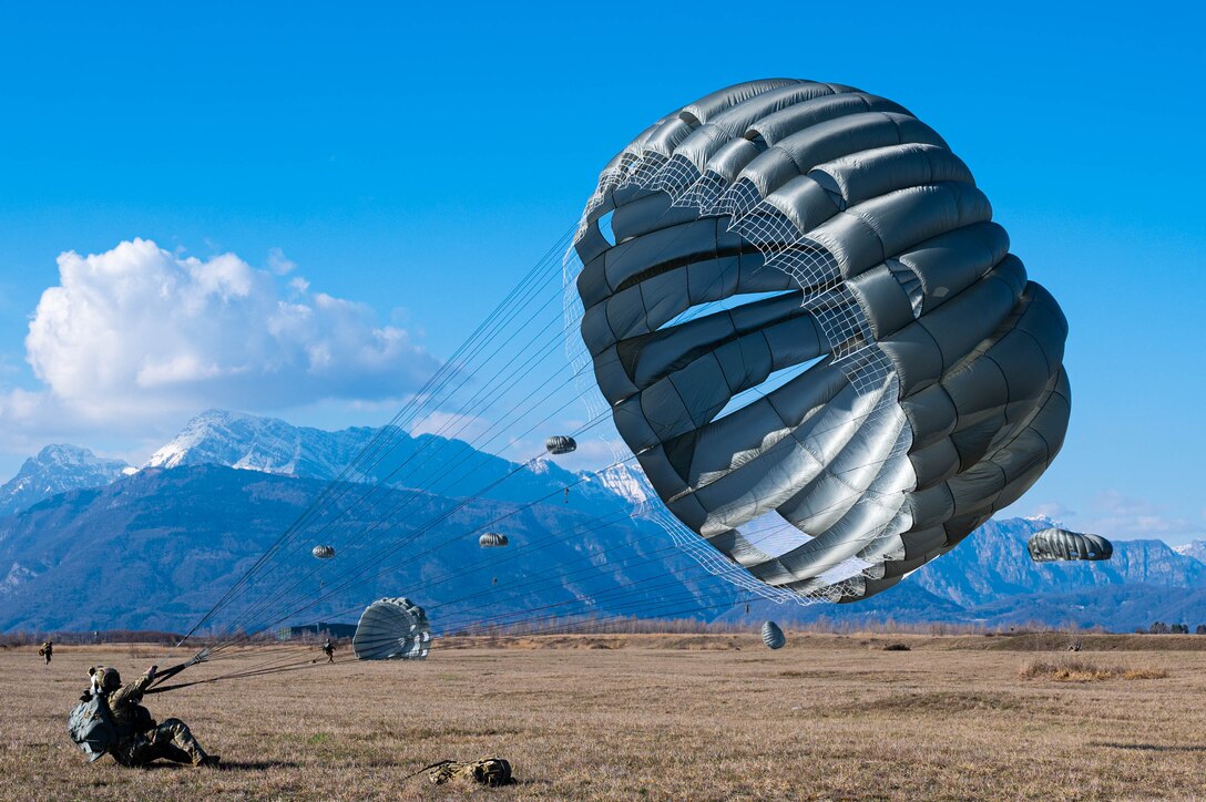 An Air Force pararescuman takes control of a gray parachute after landing with other parachutes and mountains in the backdrop.