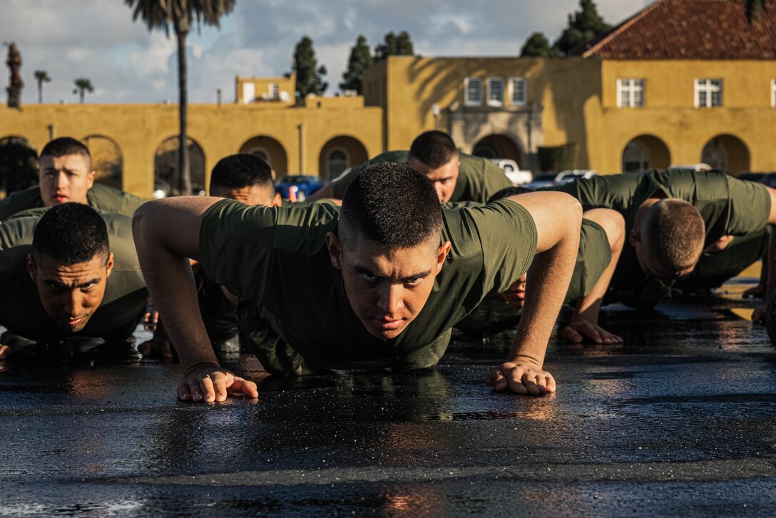 Several Marines perform pushups with yellow buildings and trees in the background.