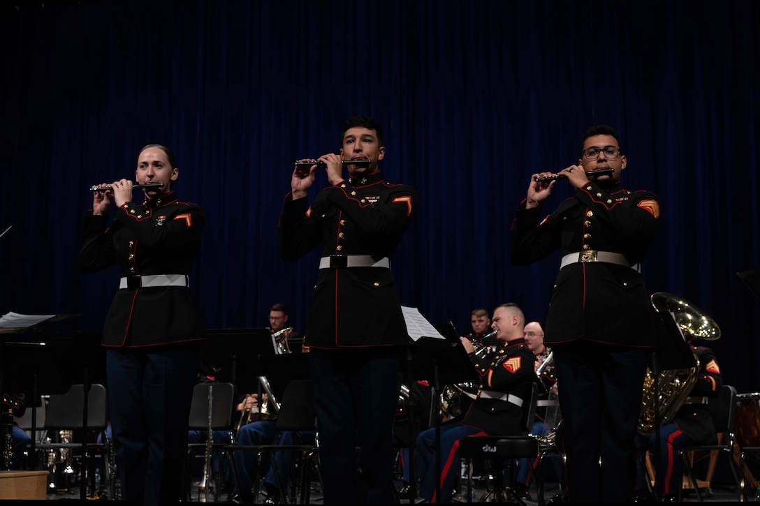 Three Marines play piccolo on a dark stage with an orchestra in the background.