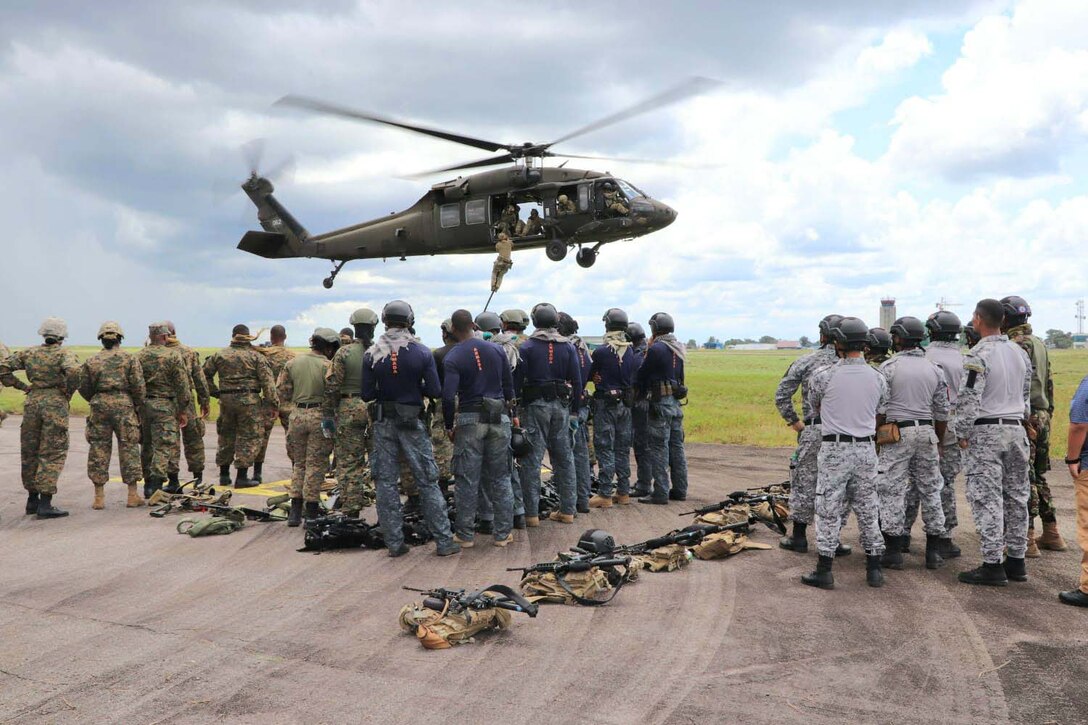 A soldier dangles from rope hanging from a hovering helicopter as others on the ground watch.