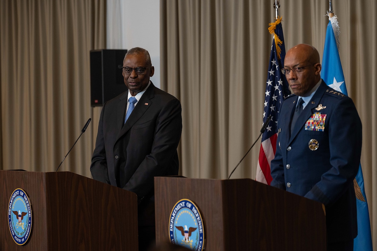 A person dressed in business attire and a military member stand behind lecterns with flags in the background.
