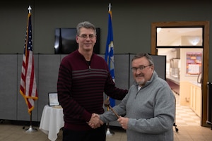 Two men pose for a photo. The one at left, in a gray sweater, hands the taller man, at right in a maroon sweater with gray and black stripes, a wooden gavel.
