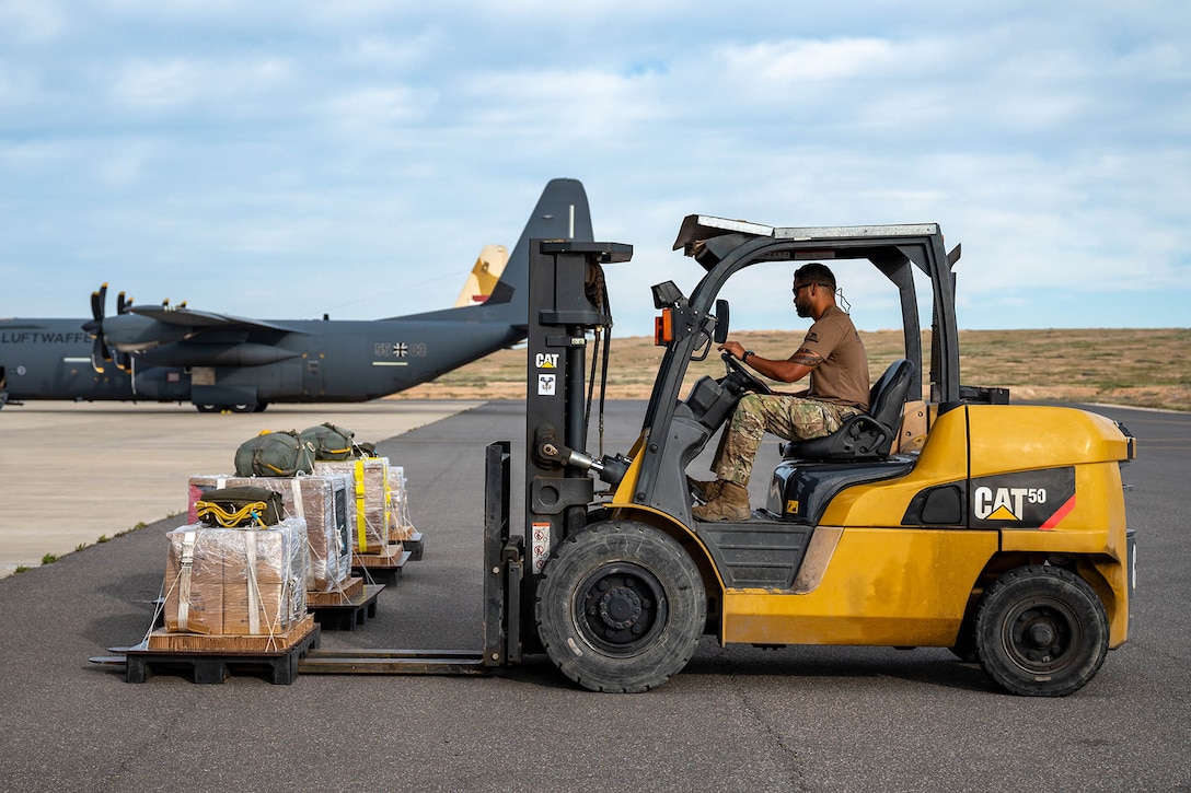 An airman operates a forklift holding cargo on a flight line, with an aircraft parked in the background.
