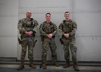 Three men in military uniform stand facing the camera against a plain wall.