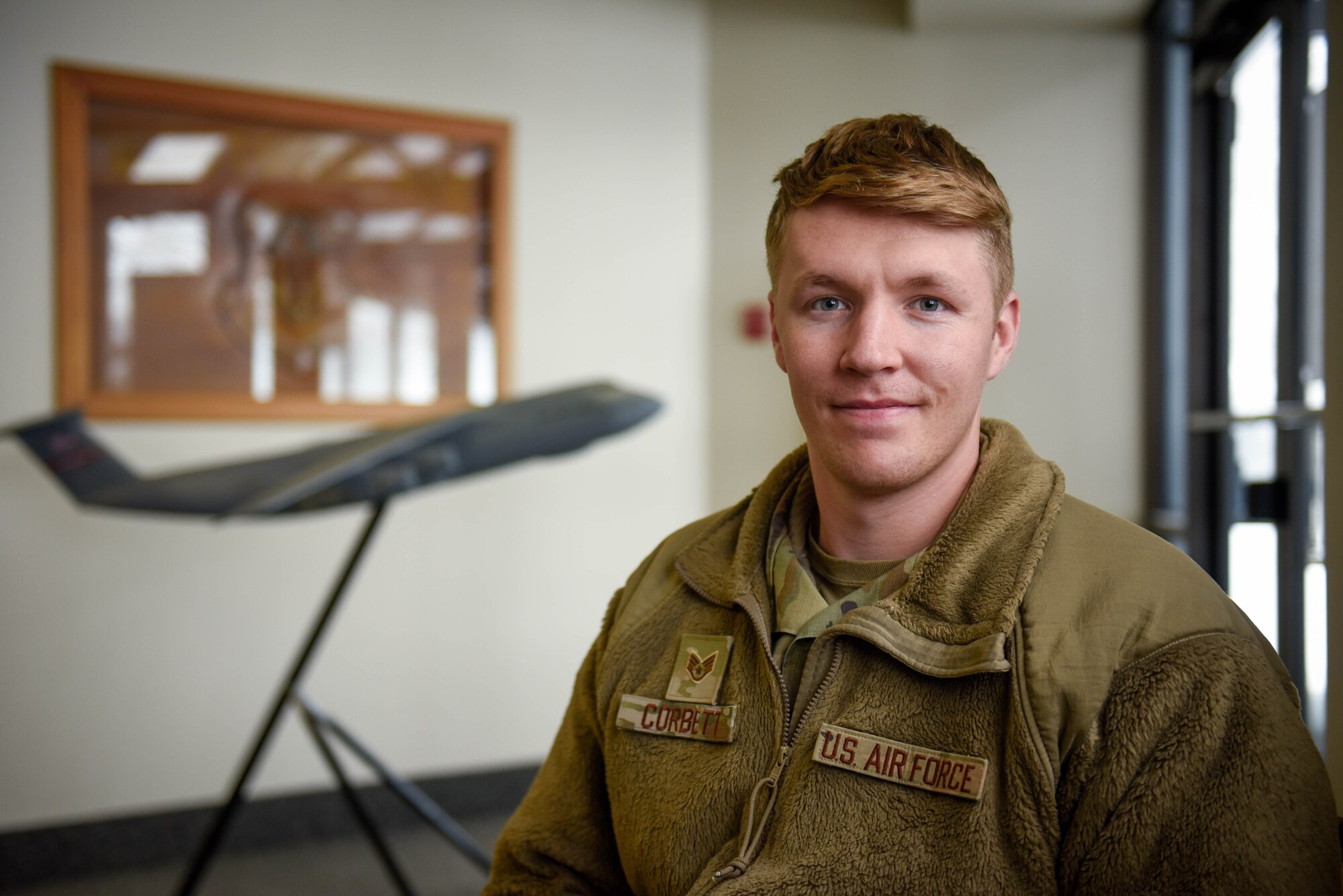 U.S. Air Force Staff Sgt. Kel Corbett, wearing his OCP uniform, poses in front of a C-5M Super Galaxy display near the base commander's office.