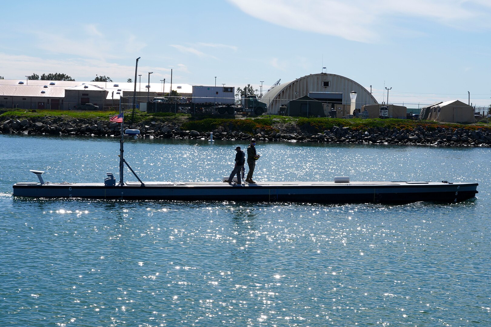 Two people stand atop a low-profile water vessel. Warehouses and other buildings are on the shore.