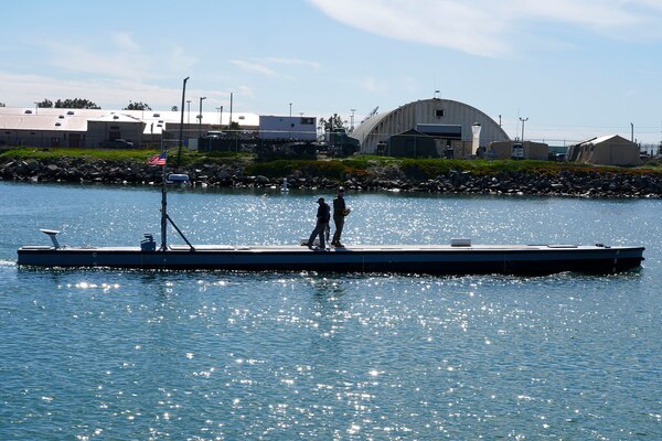 Two people stand atop a low-profile water vessel. Warehouses and other buildings are on the shore.