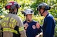 Two people with dark blue shirts and multicolored protective helmets are talking while someone dressed in firefighter clothing (large beige coat with bright yellow reflective stripes and the words Technical Rescue and a red helmet) is standing nearby listening>