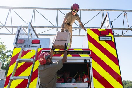 Two people wearing light brown shirts and khaki pants are handing each other a tan-colored plastic container on the backside of a fire engine that is painted with bright red and yellow diagonal stripes.