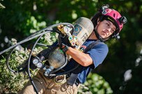 Woman with dark blue shirt is carrying a large metal tank in front of her body that has several black hoses around it. She has a red helmet on her head.