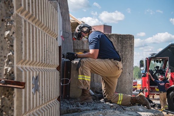 Person wearing a dark blue shirt and khaki work pants with bright yellow stripes near the calves of the pants is kneeling on one knee while working on a large piece of metal next to a large grey concrete wall. There are other similarly dress people and machinery in the background.