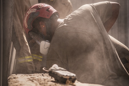 Someone dressed in khaki work pants and a light brown t-shirt is working on something around large slabs of grey concrete. He has a red helmet on with a clear facemask, and there is dust in the air.