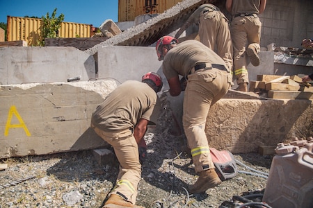Several people dressing khaki work pants and light brown t-shirts are working on something around large slabs of grey concrete. The have red helmets on.