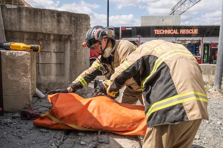 Two people in light and dark brown coats with bright yellow reflective bands around the waist and chest are either wrapping up or unwrapping a bright orange cloth that is resting on a cement ledge with debris scattered around. In the background is a truck with the words Technical Rescue.