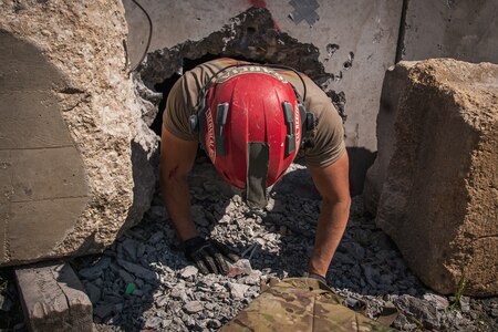 A man with a red helmet and light brown t-shirt is crawling out from a hole that was drilled into a large slab of grey concrete. There is lots of rubble around him.