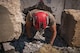 A man with a red helmet and light brown t-shirt is crawling out from a hole that was drilled into a large slab of grey concrete. There is lots of rubble around him.