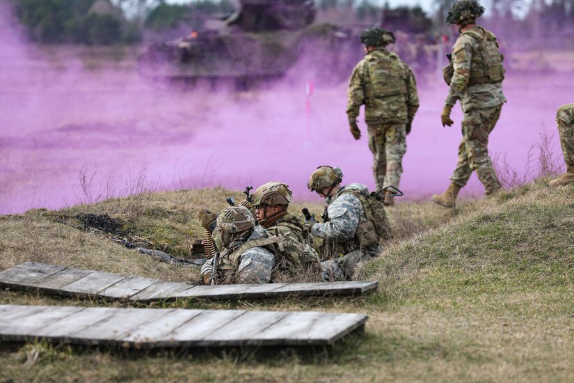 Three soldiers stand behind a grassy hill while two others walk near them as purple smoke is before them.