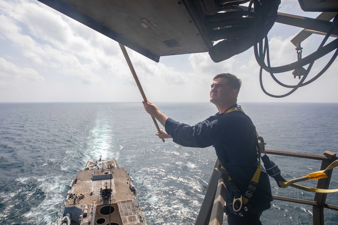 A sailor adjusts an antenna while strapped to a lift with water and a ship below.