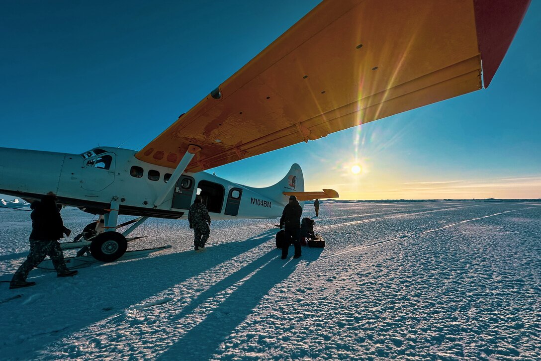 A small aircraft with orange wings is parked on a snowy runway with three troops standing next to it and a bright sun in the background.