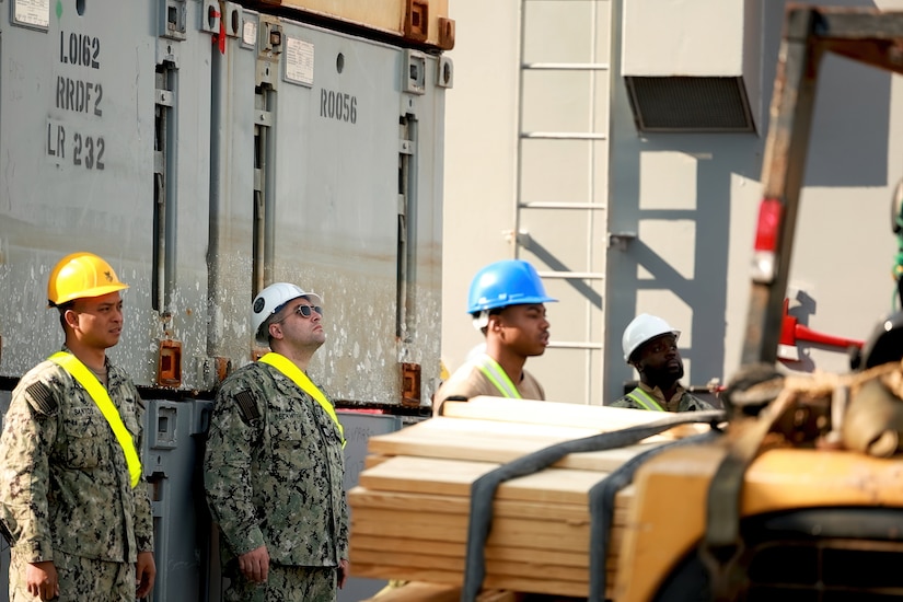 Service members in uniform and wearing hard hats stand near equipment on a ship.