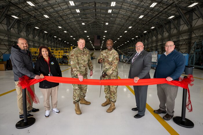 Members pose for a group photo from behind a red ceremonial ribbon while Maj. Gen. Bell prepares to cut the ribbon with large scissors.