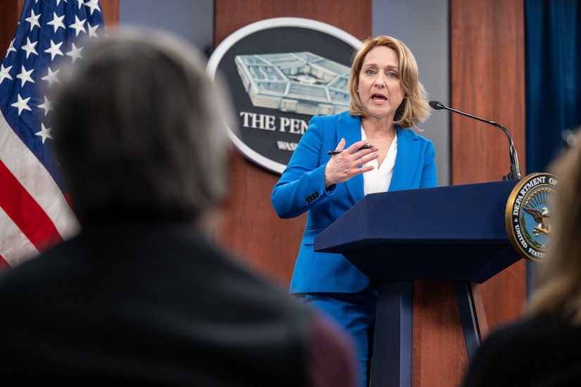 A woman standing at a lectern speaks to a group of people. The sign behind her indicates that she is at the Pentagon.