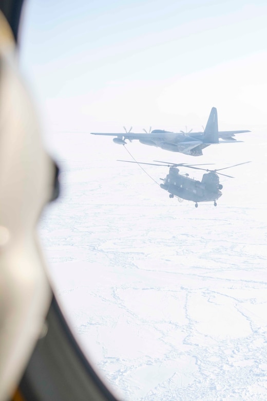 An aircraft extends its boom to a helicopter while flying over a frozen body of water as seen through a window of another aircraft.