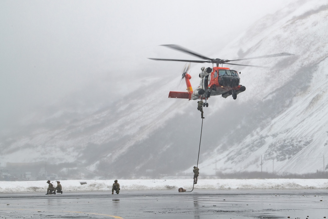 Service members rappel down a rope attached to a hovering helicopter with a snowy mountain in the background.