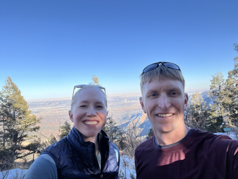 Two soldiers pose for a photo and smile with trees and a mountain landscape in the backdrop.