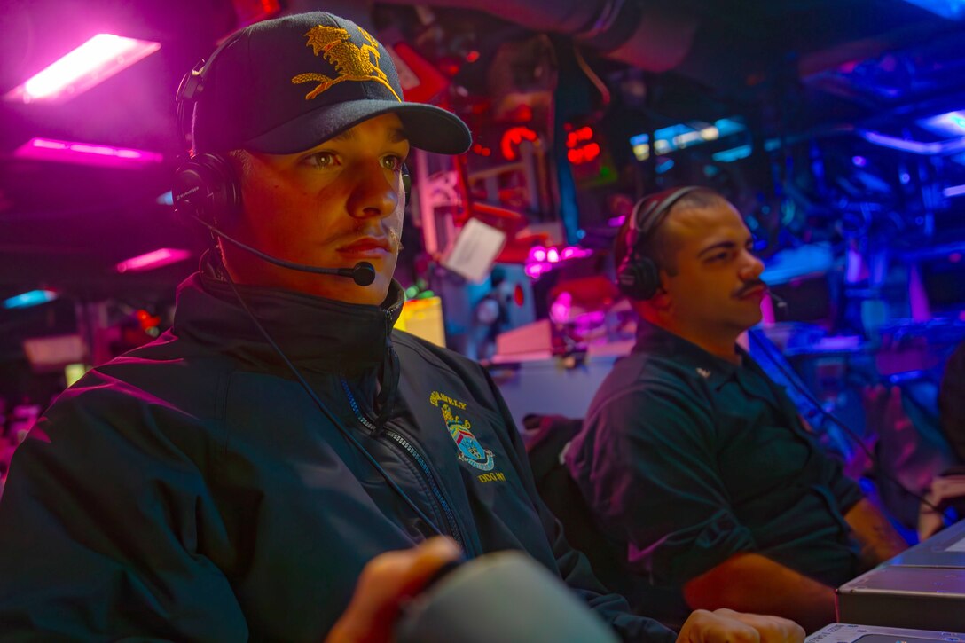 Two sailors sit in front of metal desks looking forward in an enclosed room with different colored lights