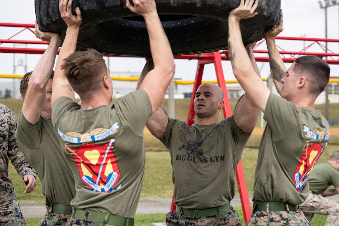 Four Marines lift a tire above their heads with red and  yellow beams in the backdrop.