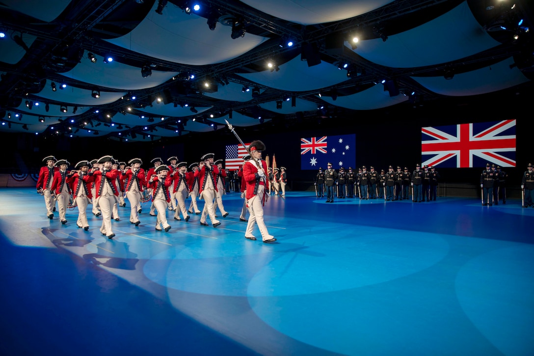 Soldiers dressed in ceremonial uniforms play instruments while moving across a stage in front of U.S., British and Australian troops standing in front of their countries’ flag.