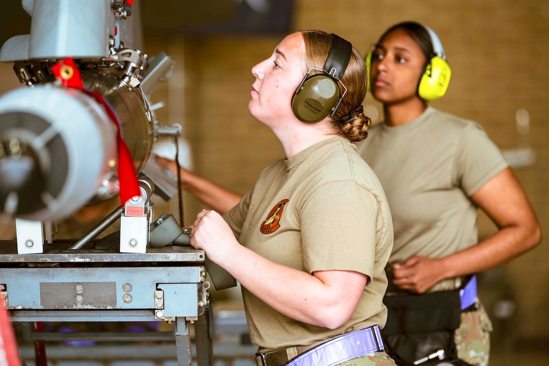 Two airmen standing next to each other work on a weapon during a competition.