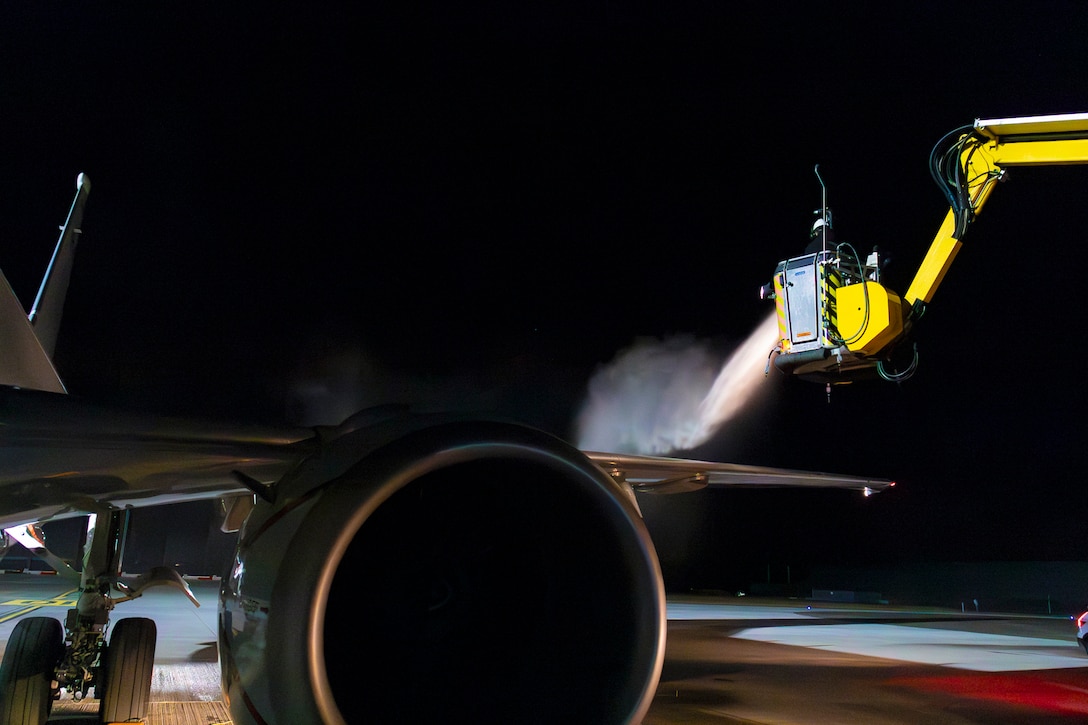 A sailor sitting in a de-icing truck sprays a chemical onto the wing of an aircraft parked on a tarmac at night.