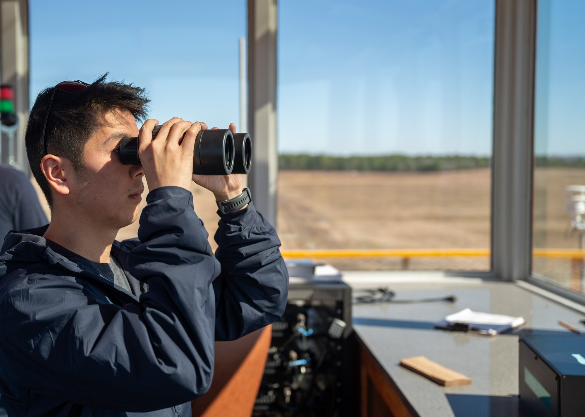 A man looks through binoculars out of an air traffic control tower.