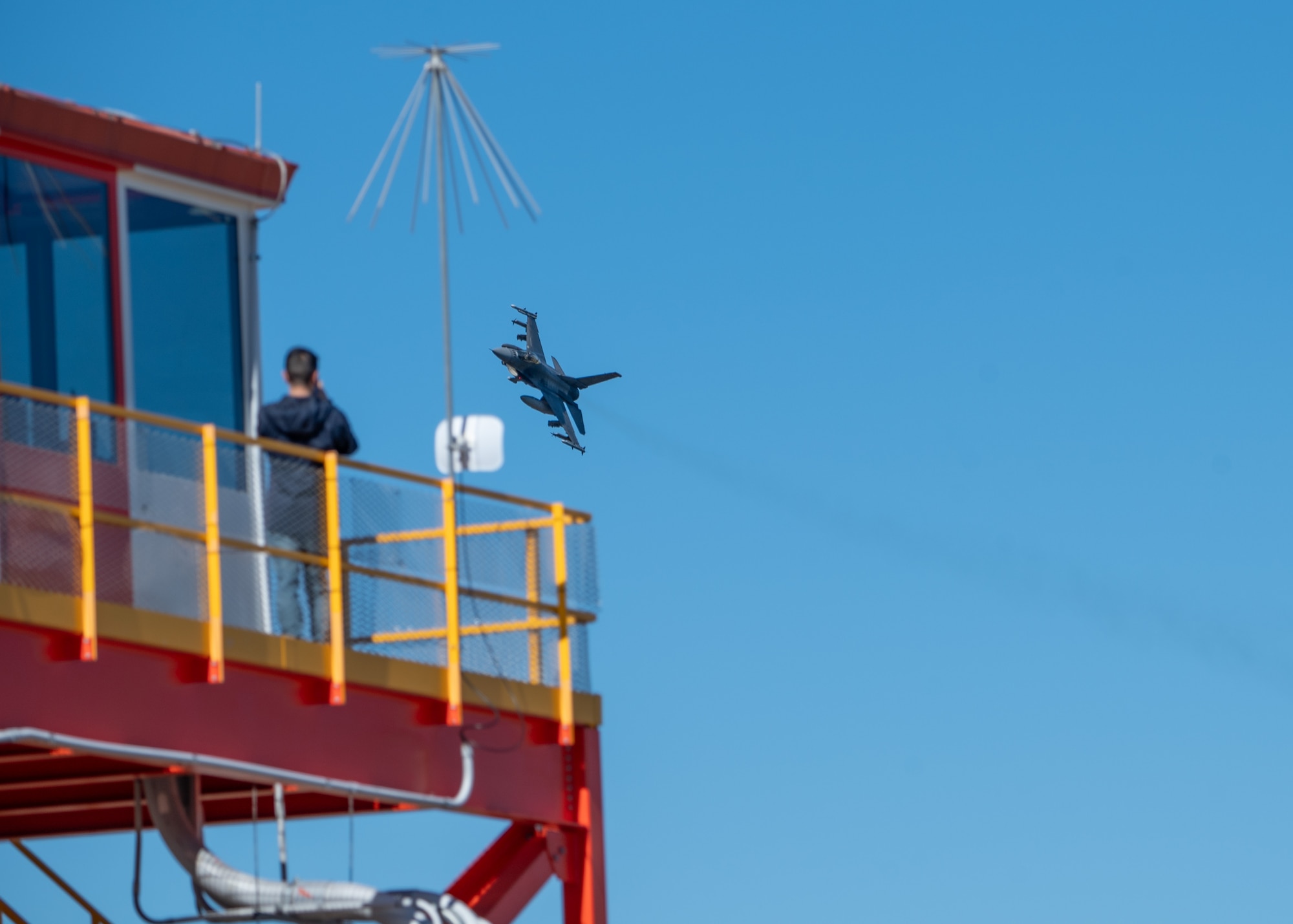 A man on an air traffic control tower stands on the balcony observing a jet as it passes behind the tower.