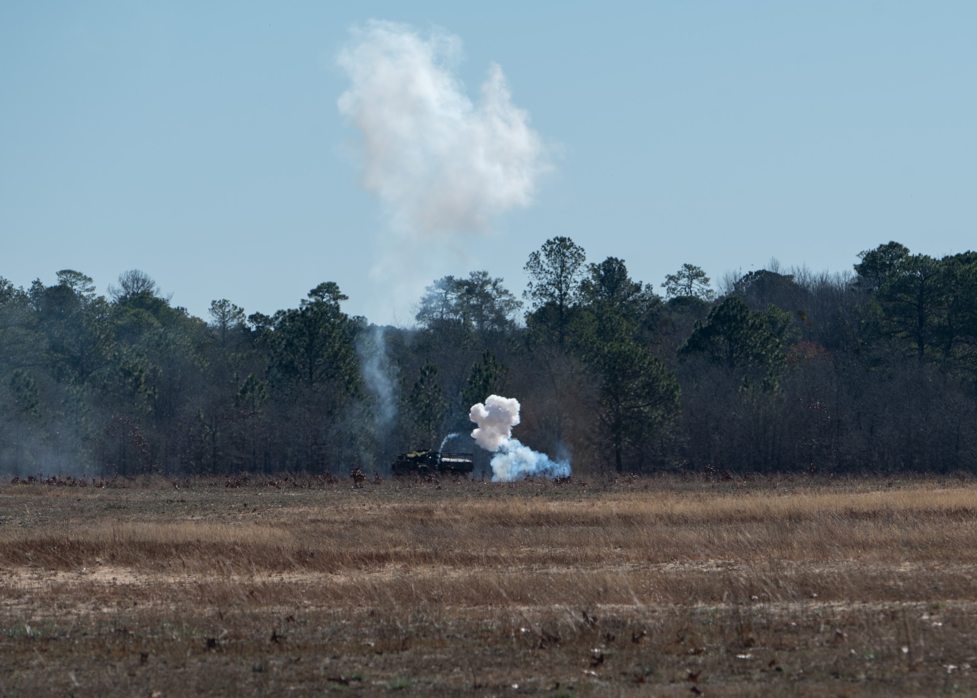 A wide shot of a field with a small plume of smoke near the ground, and a taller plume of smoke up in the air.