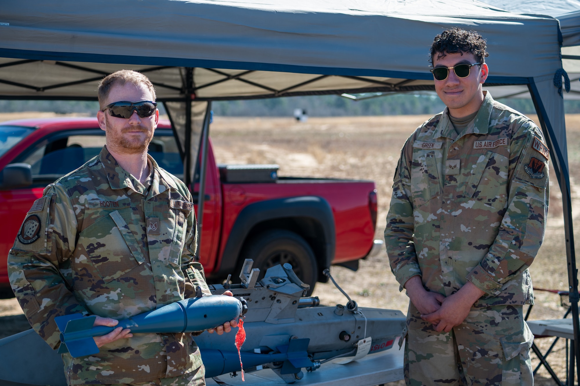 Two men stand side-by-side for a photo, the man on the left i holding a small munition - the BDU-33.
