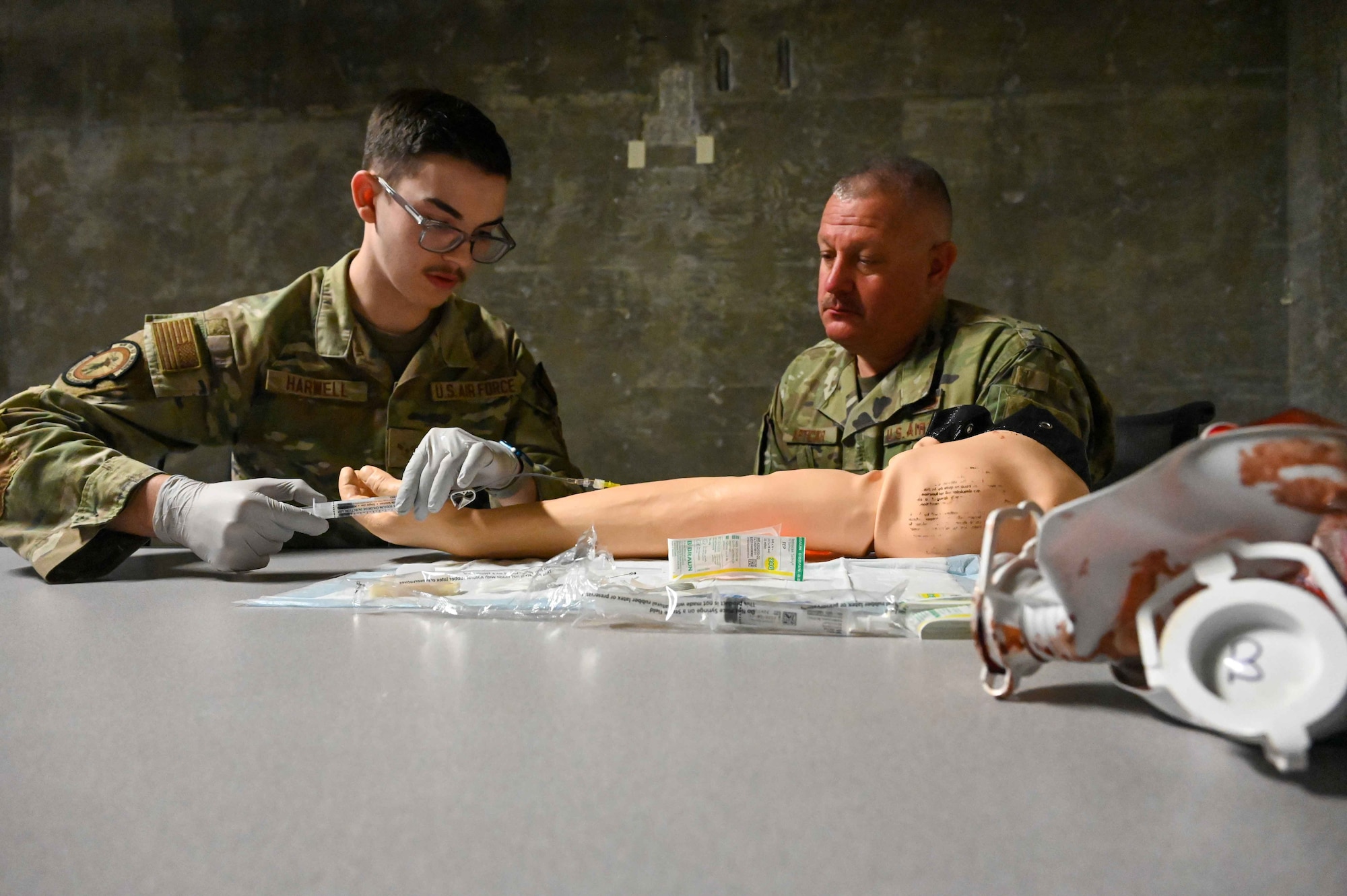 U.S. Air Force Airman 1st Class Mason Harwell, left, 97th Medical Group (MDG) aerospace medical technician, demonstrates to Chief Master Sgt. Justin Apticar, 19th Air Force command chief, right, how to insert an intravenous injection at Altus Air Force Base, Oklahoma, March 12, 2024. While at the 97th MDG, Apticar also learned how to create a dental mold. (U.S. Air Force photo by Airman 1st Class Kari Degraffenreed)