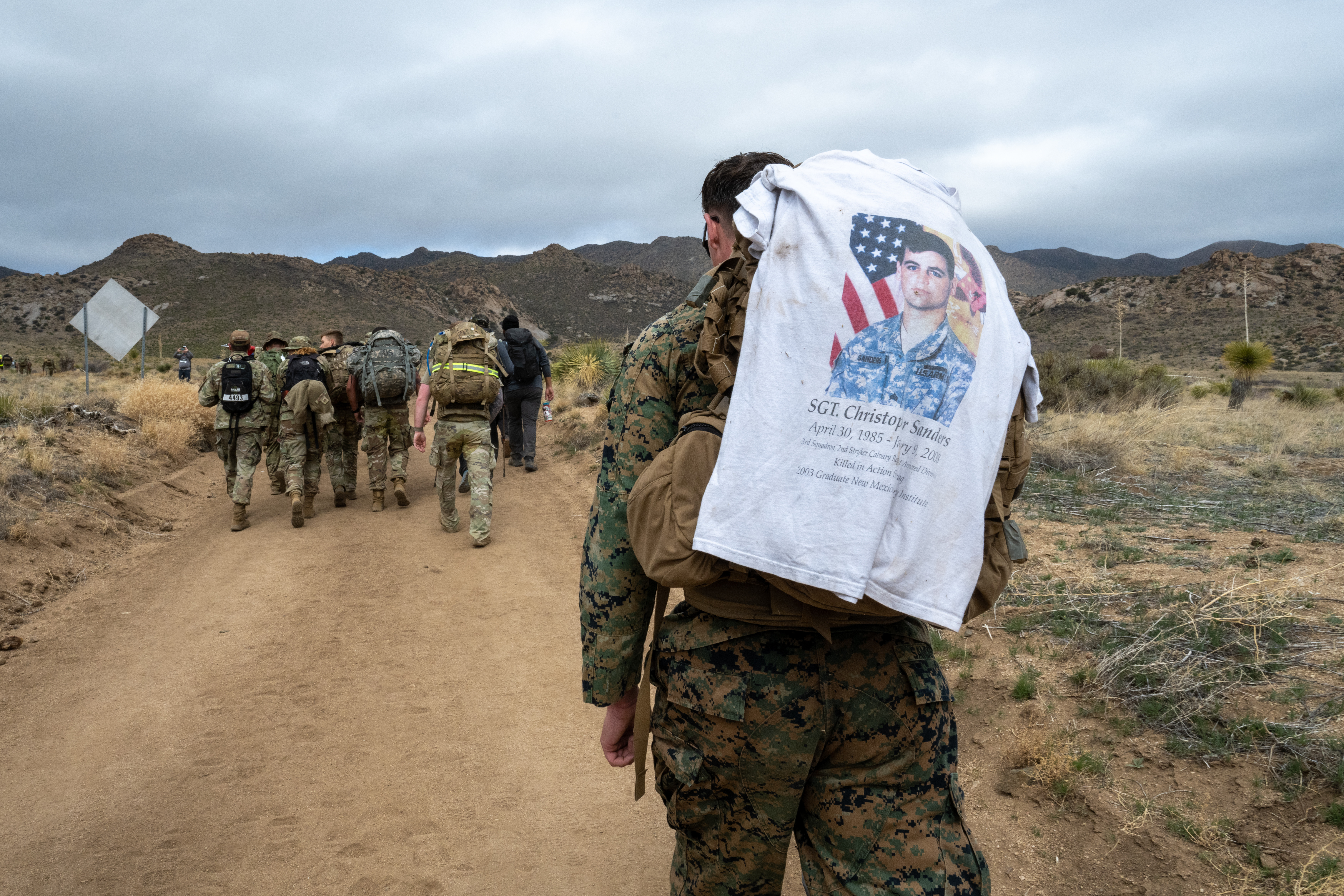 A service member in uniform walks along a dirt path in the desert with a T-shirt draped over a rucksack on his back, while others walk ahead.