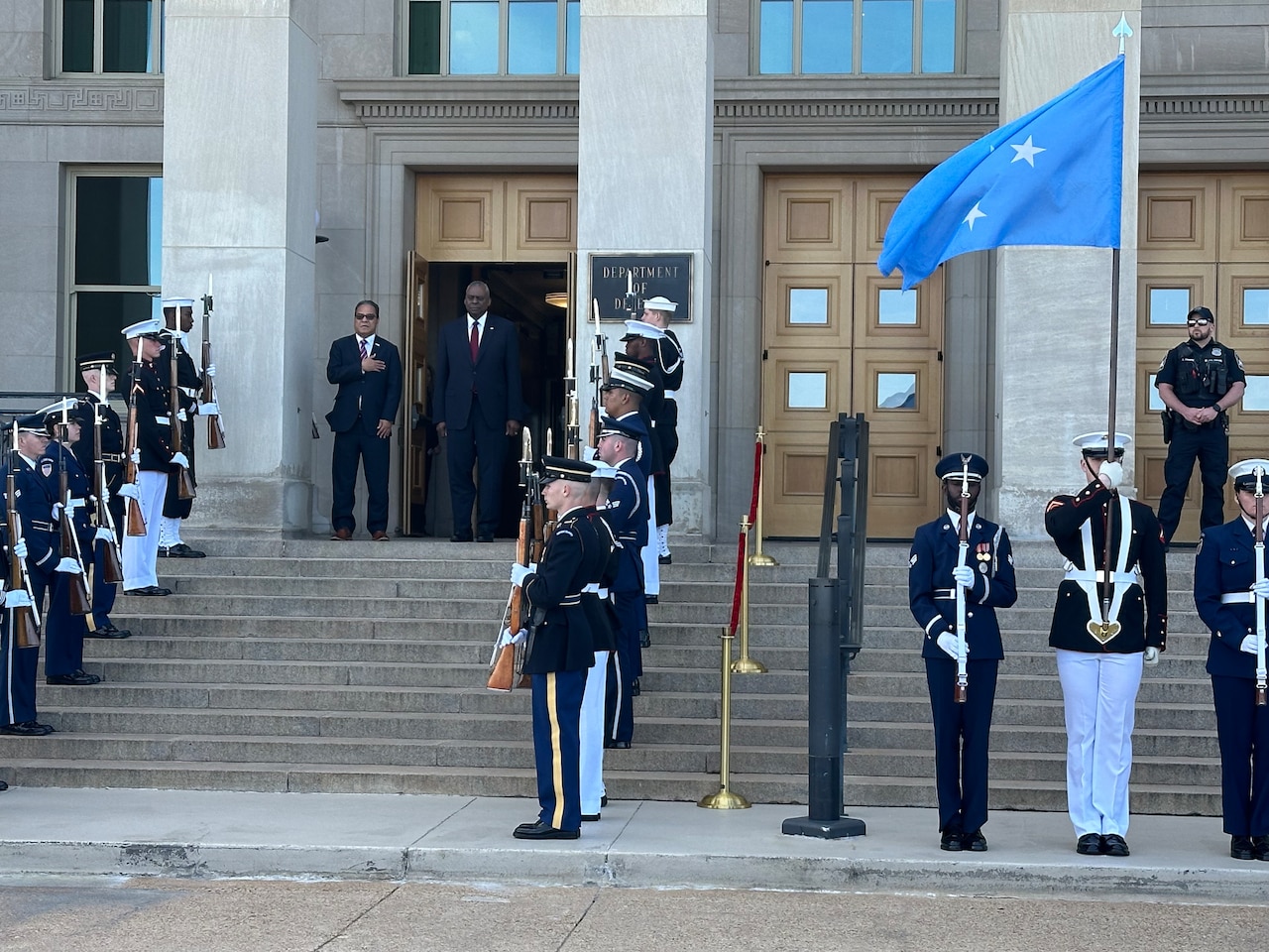 Two civilians stand on steps while flanked by a ceremonial detail of U.S. service members.