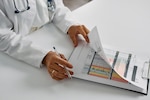 Provider wearing white lab coat flips through papers with charts on clipboard.