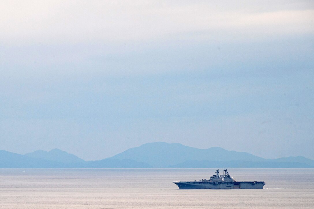 A military ship steams across the water with hazy mountains in the background.