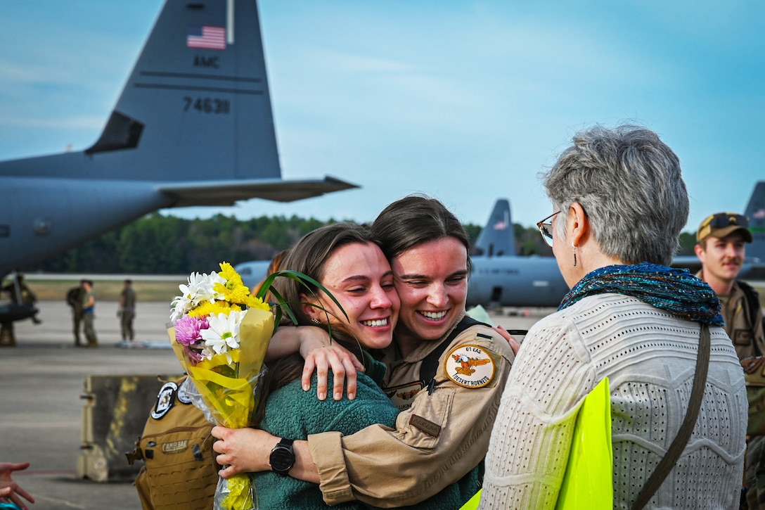An airman holds a bouquet of flowers while getting hugged by a civilian. Another civilian stands close by and watches.