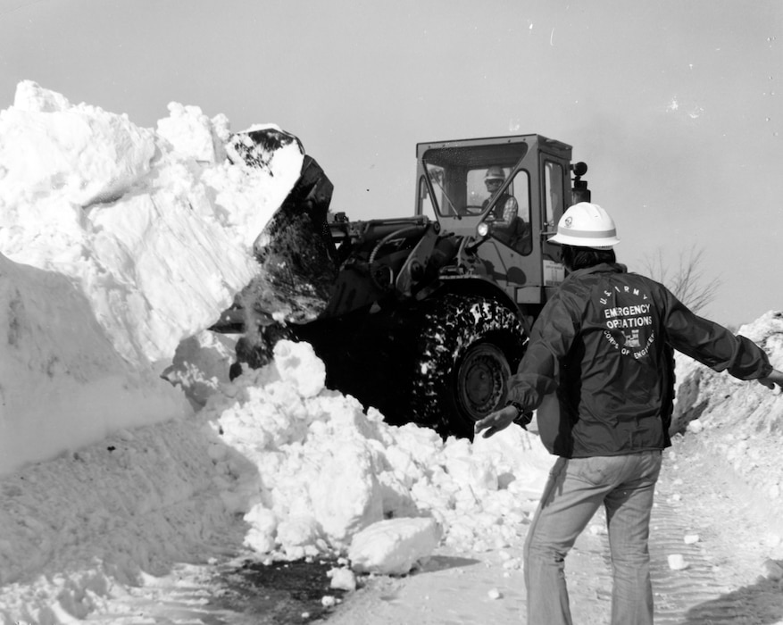 Man in USACE jacket observes large equipment shoveling snow.