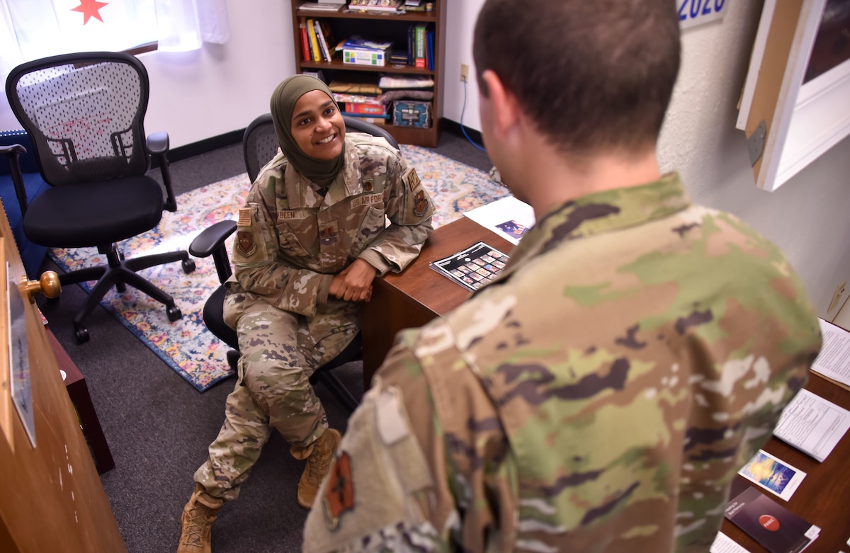 A woman in a military uniform sitting down at a desk, wearing a hijab is talking to a student airman in her office.