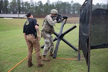 Man in USAMU shooting uniform instructing U.S. Army Soldier on outdoor firing range.